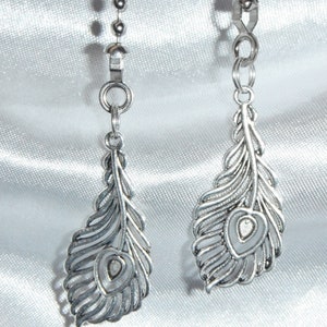 May include: Two silver-toned feather-shaped ceiling fan pull chains. Each chain features a detailed feather pendant with a central heart-shaped design. The chains hang from a silver ball chain against a white satin background.
