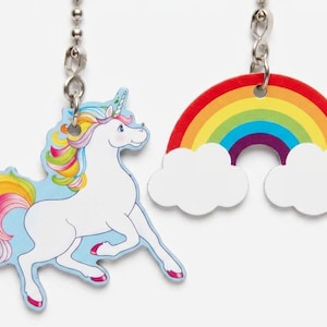 May include: Two whimsical ceiling fan pulls. One is a white unicorn with a colorful mane and tail, set against a light blue background. The other is a rainbow with white clouds. Both are attached to silver chains.