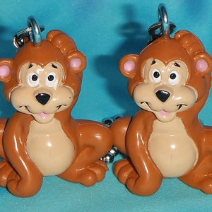 May include: Two brown bear keychains with pink ears and black noses. The bears are sitting with their paws tucked in and have a smiling expression.