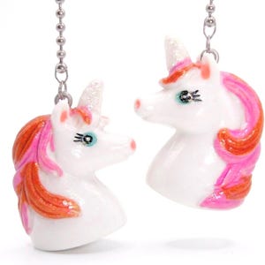 May include: Two white unicorn head pendants attached to silver-toned metal ball chains. Each unicorn features a glittery white horn, a bright pink and orange mane, and painted eyes with long black eyelashes. The pendants are suspended vertically.