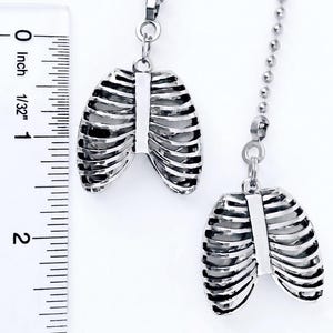 May include: Two silver-colored pendant necklaces, each featuring a detailed rib cage design. The pendants are suspended from delicate chains. A ruler is placed next to the pendants, showing measurements in inches. The pendants are a unique accessory.