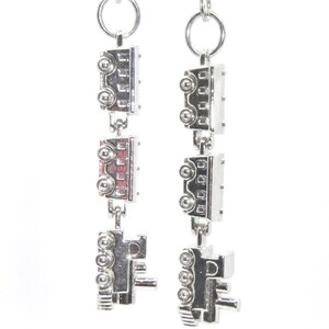 May include: Silver-toned dangle earrings. Each earring features three connected rectangular charms with circular and square details. The earrings hang from a small silver ring and a hook. The design has a mechanical or industrial aesthetic.