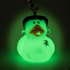 May include: A glow-in-the-dark rubber duck charm featuring a black hairpiece, bolts on the sides of the head, and a stitched scar detail on the chest. The duck has a dark orange beak and is attached to a silver-toned metal ball chain connector.