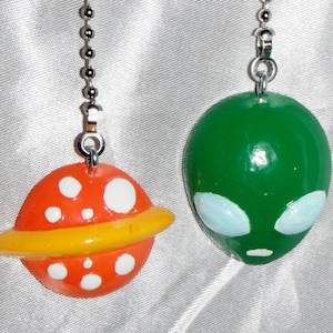 May include: Two whimsical ceiling fan pulls. One is an orange planet with white spots and a yellow ring. The other is a green alien head with light blue eyes and a small mouth. Both are attached to silver ball chains.