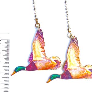 May include: Two decorative duck-shaped ceiling fan pulls. Each pull features a flying duck with orange, purple, and green coloring. The pulls are attached to a silver ball chain. The ruler shows the pulls are approximately 2 inches long.