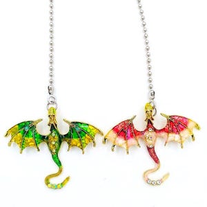 May include: Two metal dragon ornaments hanging from silver ball chains. One dragon features green and yellow enamel wings with small clear crystals, while the other has red, pink, and white enamel wings with clear crystals along its body and tail.