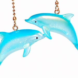 May include: Two light blue dolphin-shaped ceiling fan pull chains. The dolphins are translucent with glitter accents and black eyes. They hang from gold-colored chains and are set against a white background.