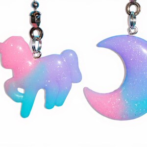 May include: Two glittery, pastel-colored charms hang from silver chains. One is a unicorn in pink, blue, and purple. The other is a crescent moon in similar hues. These charms are likely for jewelry or decorative purposes.