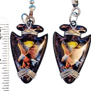 May include: A pair of arrowhead-shaped earrings. Each earring features a detailed image of an eagle with outstretched wings against a dark background. The earrings are approximately 1 inch long and have silver-colored wire accents.
