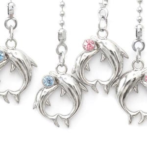 May include: Silver dolphin pendants with blue and pink crystal accents. Each pendant features two dolphins forming a heart shape. The pendants are attached to a silver chain.