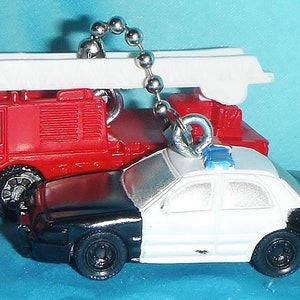 Könnte beinhalten: Ein roter Feuerwehrauto- und ein schwarz-weißes Polizeiauto-Schlüsselanhänger. Der Feuerwehrauto hat eine weiße Leiter oben. Das Polizeiauto hat ein blaues Licht. Beide sind an einer silbernen Kugelkette befestigt.