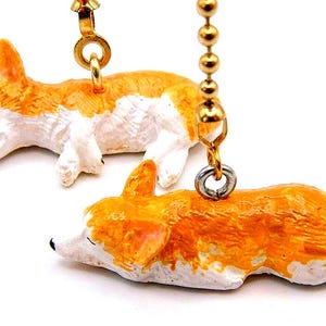 May include: Two orange and white corgi dog figurines, attached to gold-colored chains. The corgis are lying down, with their eyes closed. The background is white.