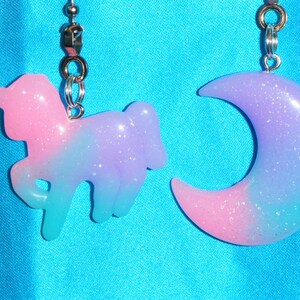 May include: A pair of earrings with a pink and blue ombre effect. One earring is shaped like a unicorn and the other is shaped like a crescent moon.