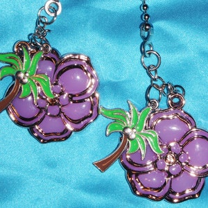 May include: Two purple flower-shaped charms with gold trim and a palm tree design. The charms are suspended from a silver chain. The background is a light blue fabric.