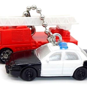 Può includere: Portachiavi in miniatura con un'autopompa rossa e un'auto della polizia bianca e nera. L'autopompa ha una scala bianca sopra. L'auto della polizia ha una luce blu sopra. Entrambi sono attaccati a una catena a sfera d'argento.
