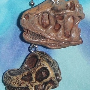 May include: Two brown dinosaur skull pendants hanging from a silver chain. The pendants are detailed with visible teeth and eye sockets, and are attached to a ball chain. The background is a light blue.