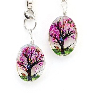 May include: A pair of oval-shaped resin pendants hanging from silver-toned metal chains. Each pendant features a detailed miniature painting of a tree with vibrant pink blossoms, a dark brown trunk, green grass at the base, and small butterflies.