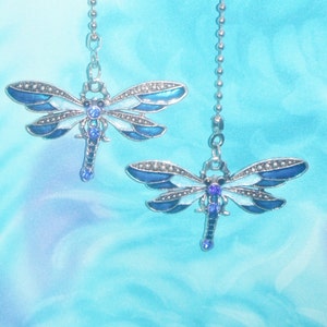 May include: Two dragonfly-shaped ceiling fan pulls. Each dragonfly is silver-toned with blue and white enamel wings, and a blue crystal accent. The pulls hang from a silver chain against a blue background.