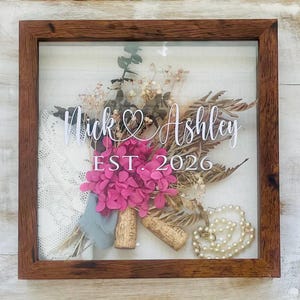 Puede incluir: Una caja de sombra enmarcada con un ramo de flores secas, incluyendo hortensias rosas. El texto "Nick & Ashley EST. 2026" está en escritura blanca. El marco es marrón y el fondo es blanco.