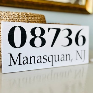 Puede incluir: Un letrero de madera blanco con texto negro. El letrero dice "08736 Manasquan, NJ".