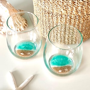 Pode incluir: Dois copos de vinho sem haste transparentes com um design de cena de praia. A base de cada copo apresenta um oceano azul turquesa, areia bege e uma pequena estrela do mar branca. Uma concha grande e uma cesta de vime estão no fundo.