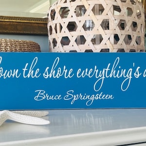 May include: A rectangular blue wooden sign featuring white cursive text that reads 'cause down the shore everything's alright' followed by the name 'Bruce Springsteen' in a smaller white font centered below the main phrase.