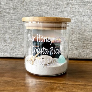May include: A clear glass jar with a bamboo lid, filled with white sand, a starfish, and a piece of sea glass. The jar contains a photo of a sunset and the words "Our Honeymoon Costa Rica July 2021".