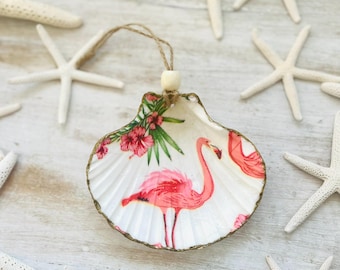May include: A pink flamingo with tropical flowers and leaves printed on a white seashell ornament. The ornament has a gold trim and a string for hanging.
