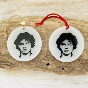 Op de afbeelding: Twee ronde, witte ornamenten met zwarte portretten. Eén heeft een natuurlijke touwhanger, de andere een rood lint. De portretten zijn van een man met donker haar en een serieuze uitdrukking. De ornamenten worden getoond op een stuk drijfhout.