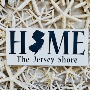 May include: White wooden sign with black lettering that reads "HOME The Jersey Shore". The sign features a black silhouette of the state of New Jersey. The sign is surrounded by white starfish.