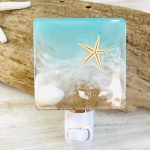 May include: Square night light featuring a beach scene with a starfish, seashells, and sand encased in resin. The top is a light blue color, and the night light has a white base with an on/off switch. The night light is a decorative item.