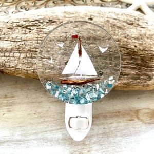 May include: A nautical-themed night light featuring a sailboat design. The clear, round light has a white sailboat with a brown hull and a red flag. Blue glass pieces represent the sea. The night light has a white plug.