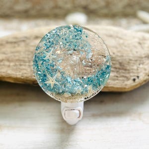 May include: A decorative night light featuring a clear, round resin design with embedded blue and white accents, small starfish, and a piece of driftwood. The night light has a white plug-in base.