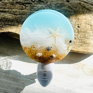 May include: A round night light with a beach scene design. The top features a light blue sky, white waves, and a sandy beach with a starfish, turtle, and seashells. The night light has a white base.