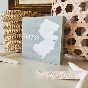 May include: A light blue wooden sign featuring a white outline of the state of New Jersey, with the zip code 08736. A heart shape is cut out of the state's outline. The sign is displayed with starfish and woven baskets.