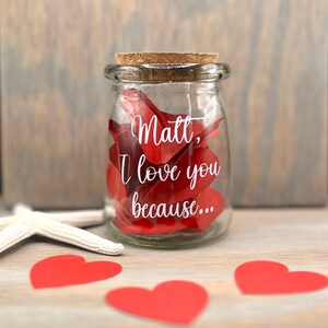 May include: A clear glass jar with a cork lid, filled with red heart-shaped paper. The jar has white text that reads "Matt, I love you because...". Three red paper hearts and a white starfish are on the wooden surface.