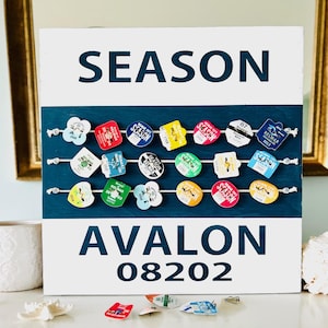 May include: A white sign with blue text that reads "SEASON" and "AVALON 08202". The sign has a blue background with rows of colourful round tags hanging from strings.