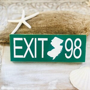 May include: A green wooden sign with the word "EXIT" and the number "98" in white, along with a white outline of the state of New Jersey. The sign is decorated with a starfish and a seashell.