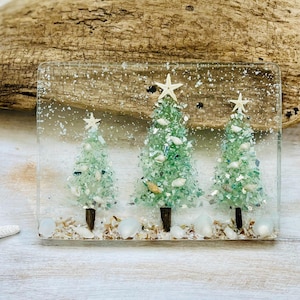 May include: A rectangular resin artwork showing three Christmas trees crafted from green and white crushed glass, each topped with a starfish. The trees are set on sand and shells, with a clear backdrop. A starfish is visible on the left.