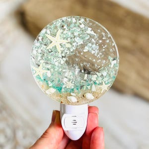 May include: A round night light with a beach scene encased in clear resin. The scene includes white starfish, sand, shells, and turquoise accents. The night light has a white base with a three-prong plug.