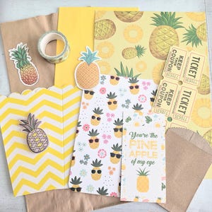May include: A collection of stationery items with a pineapple theme. Includes stickers, patterned paper, a roll of washi tape, and paper bags. One card reads "You're the PINE APPLE of my eye."