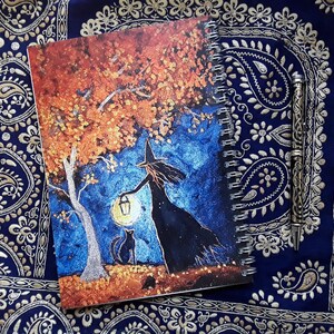 May include: Spiral-bound notebook featuring a whimsical illustration of a witch holding a lantern under a tree with a black cat. The artwork is set against a blue and orange background. A decorative pen rests beside the notebook.