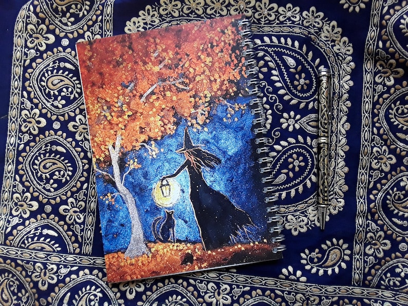 May include: Spiral-bound notebook featuring a whimsical illustration of a witch holding a lantern under a tree with a black cat. The artwork is set against a blue and orange background. A decorative pen rests beside the notebook.