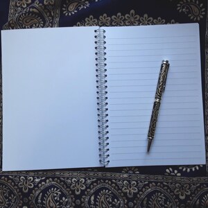 May include: An open, lined notebook with a silver spiral binding and a decorative pen. The pen has an ornate design and is resting on the right side of the open notebook. The notebook is on a dark blue and gold patterned surface.