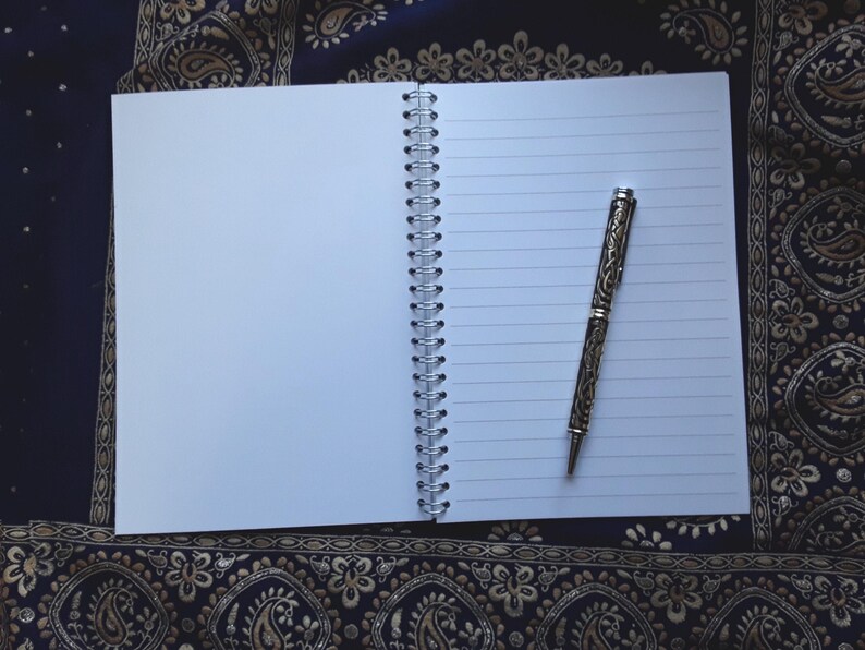 May include: An open, lined notebook with a silver spiral binding and a decorative pen. The pen has an ornate design and is resting on the right side of the open notebook. The notebook is on a dark blue and gold patterned surface.