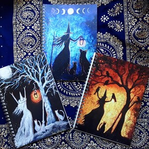 May include: Three spiral-bound notebooks with witch-themed illustrations. One features a witch with a lantern under a moonlit sky, another shows a witch and cats under a tree, and the third depicts a witch with crows under a sunset.