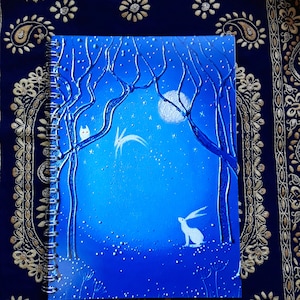 May include: A spiral-bound notebook with a blue cover featuring a whimsical forest scene. The design includes a white rabbit, an owl, a full moon, and stylized trees. The cover has a textured, embossed appearance.