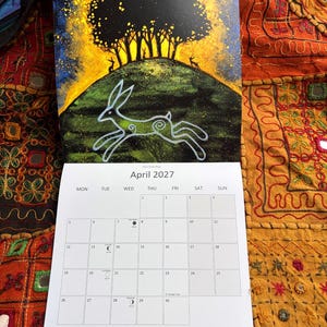 Puede incluir: Un calendario de pared para abril de 2027 con una ilustraci&oacute;n de una liebre blanca corriendo sobre una colina verde bajo un &aacute;rbol oscuro y un cielo amarillo brillante. La cuadr&iacute;cula del calendario muestra los d&iacute;as con fases lunares.