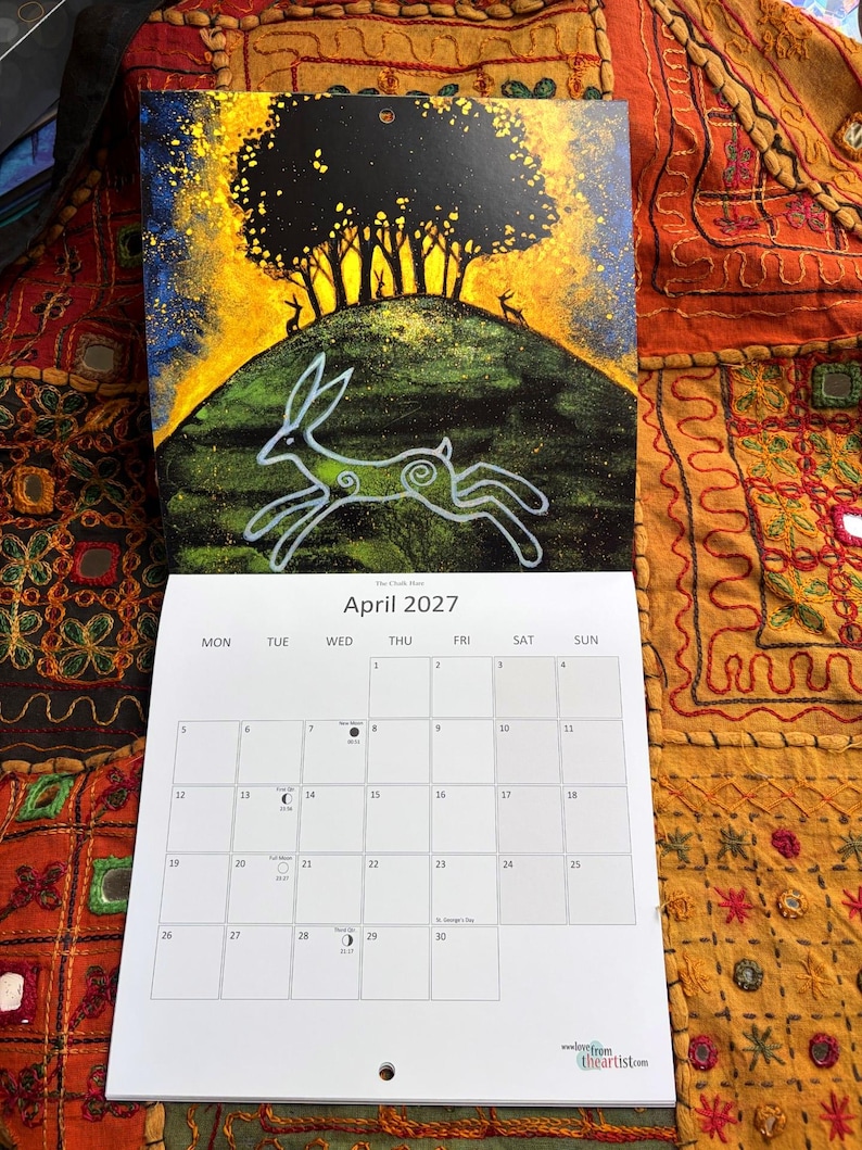 Puede incluir: Un calendario de pared para abril de 2027 con una ilustraci&oacute;n de una liebre blanca corriendo sobre una colina verde bajo un &aacute;rbol oscuro y un cielo amarillo brillante. La cuadr&iacute;cula del calendario muestra los d&iacute;as con fases lunares.