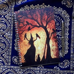 May include: Spiral-bound notebook featuring a witch silhouette with crows and a cat under a tree against a sunset backdrop. A decorative pen rests beside the notebook on a blue patterned fabric.